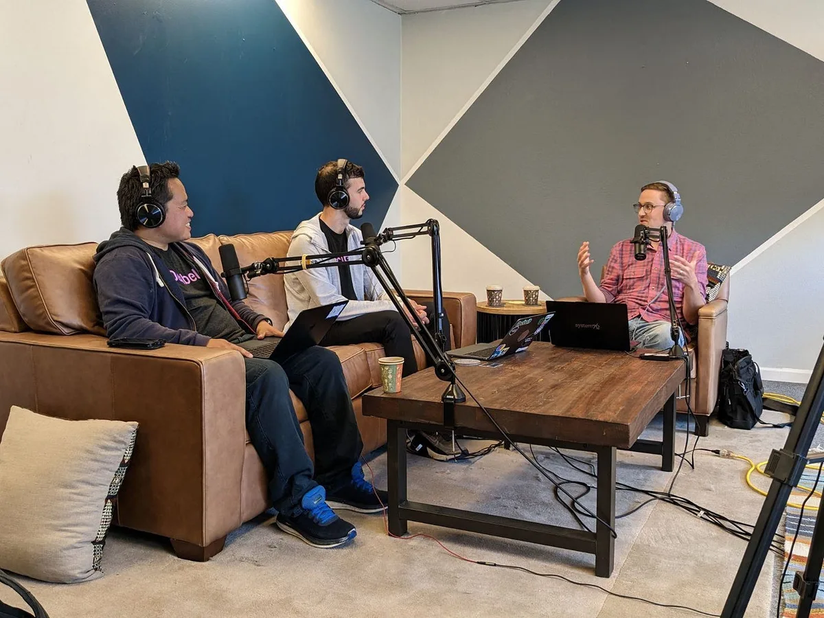 Two participants sit on a leather sofa facing a third in an armchair, with boom microphones and laptops around a wooden table.