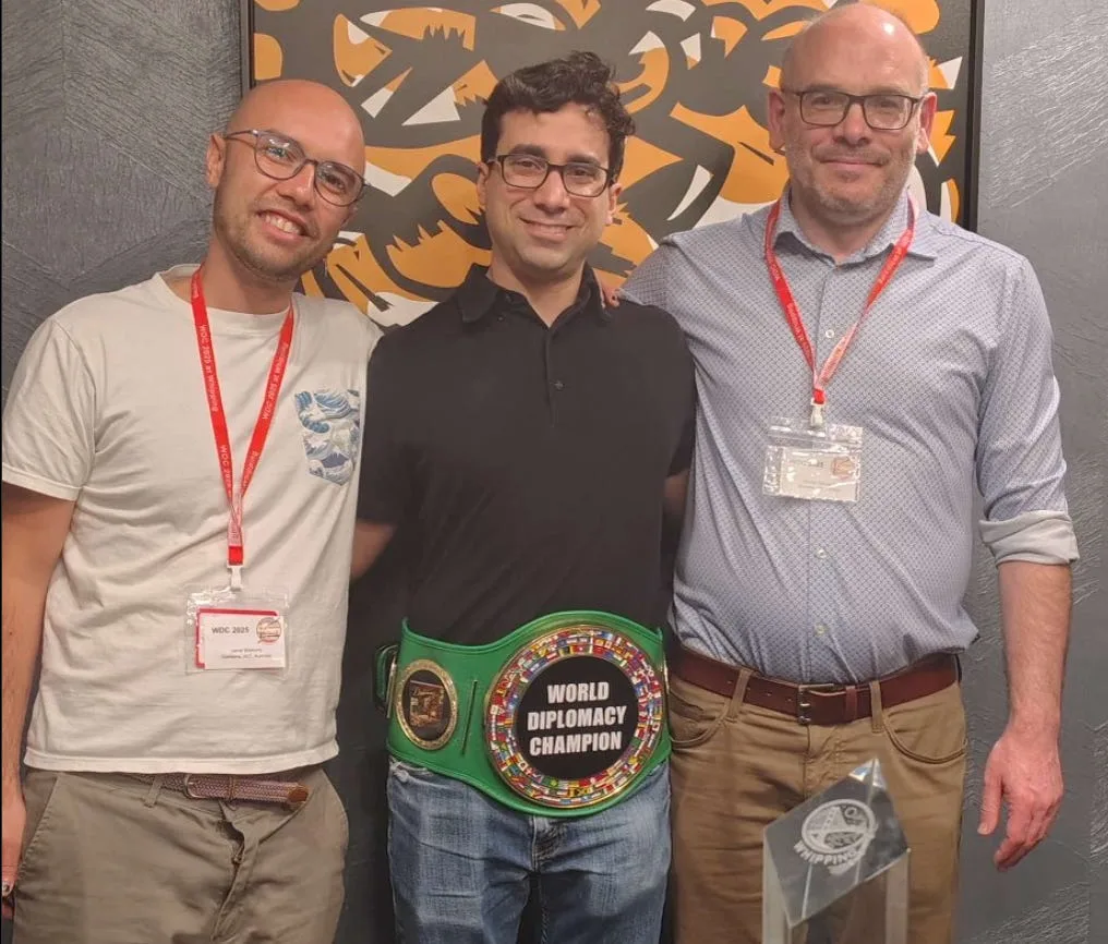 Three people standing beside one another, with a green World Diplomacy Champion belt around the center person's waist and a clear trophy in the foreground.