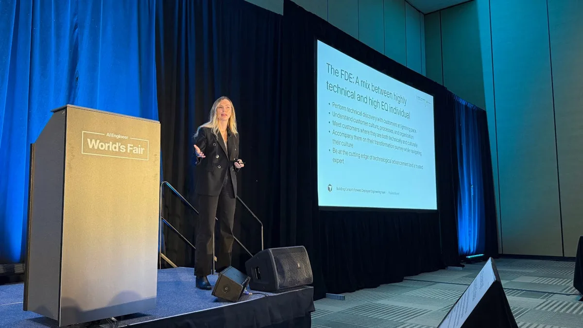 Speaker in a black suit beside a World’s Fair lectern and a projected slide about technical discovery, customer culture, and the FDE role.