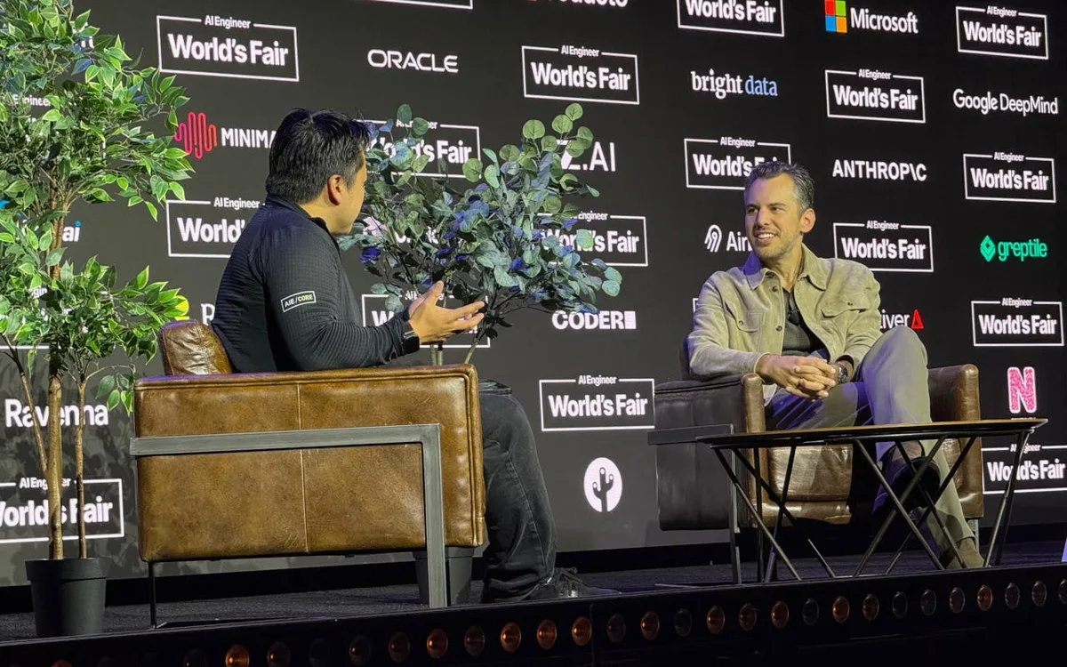 Two speakers face each other in leather armchairs on a stage backed by AI Engineer World’s Fair and sponsor signage; the speaker on the left gestures while the speaker on the right smiles.