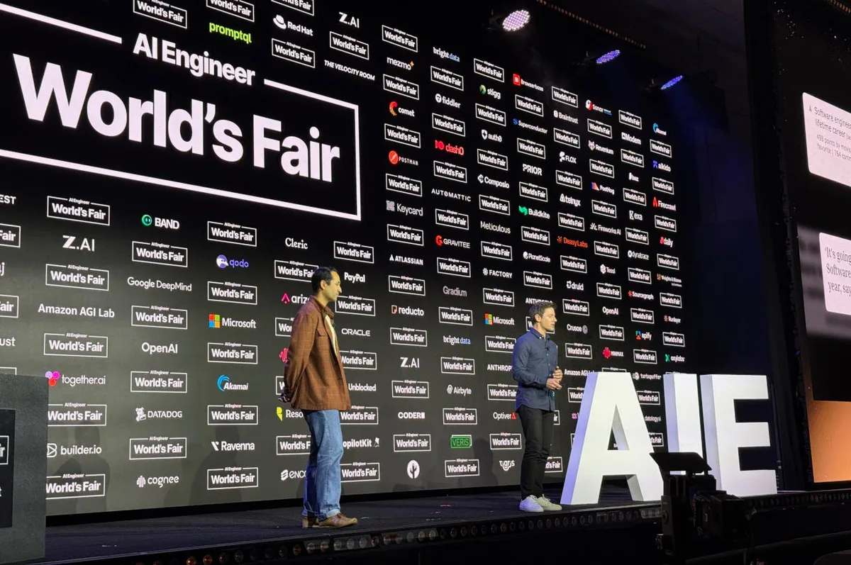 Two presenters stand on a dark stage beside large white AIE letters, with an AI Engineer World’s Fair backdrop behind them.