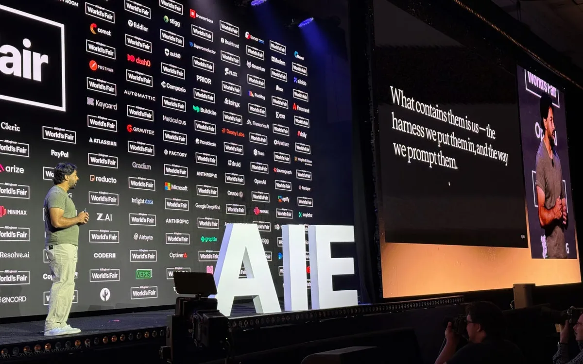 Conference speaker onstage beside large AIE letters and a projected quotation about harnesses and prompting.
