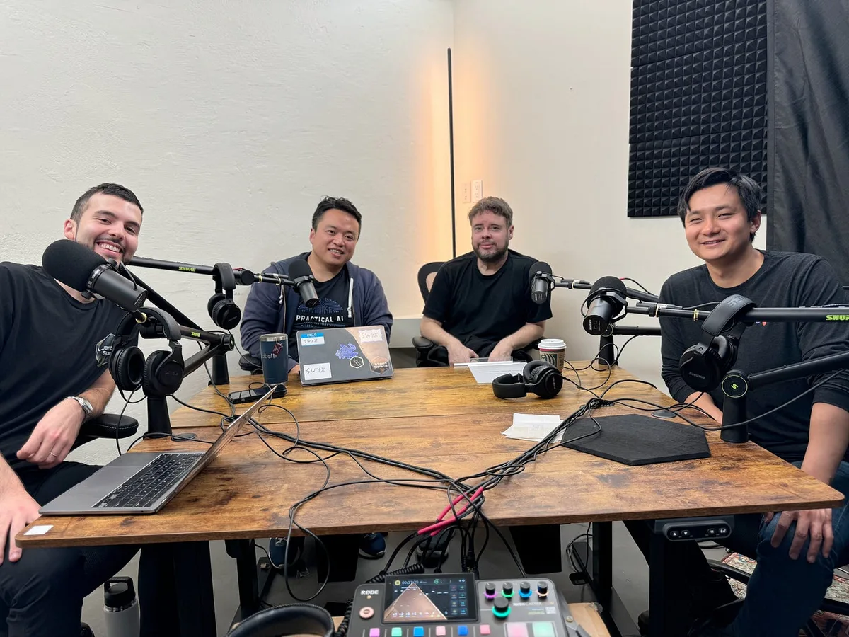 Four people seated around a wooden podcast table with boom microphones, headphones, laptops and an audio mixer.